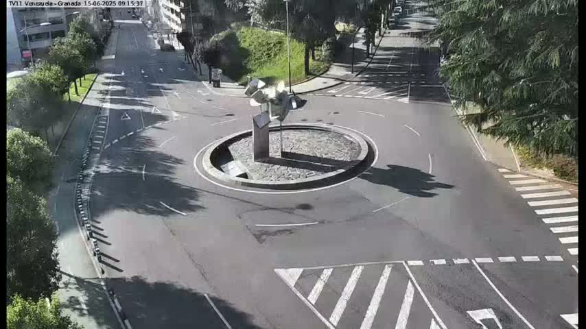 A sunny day reveals a circular traffic island with a modern sculpture in the center of a roadway intersection.