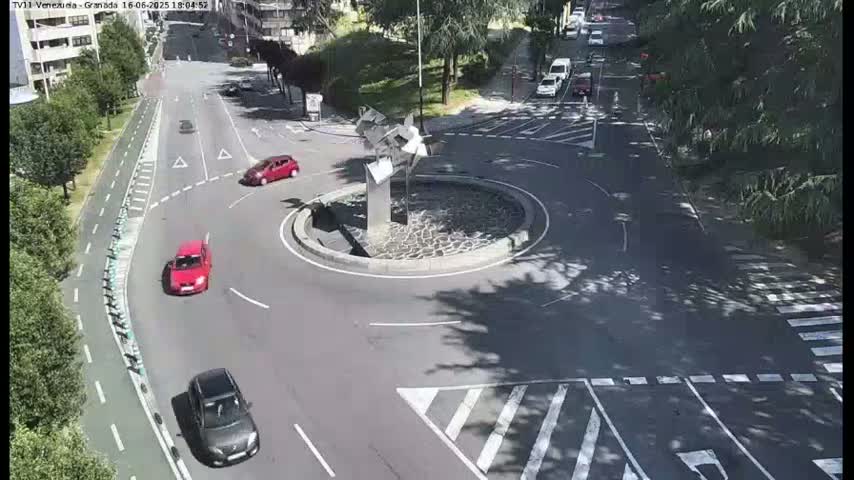 A sunny day overhead shows several cars navigating a roundabout centered by a modern metallic sculpture, surrounded by trees and buildings.