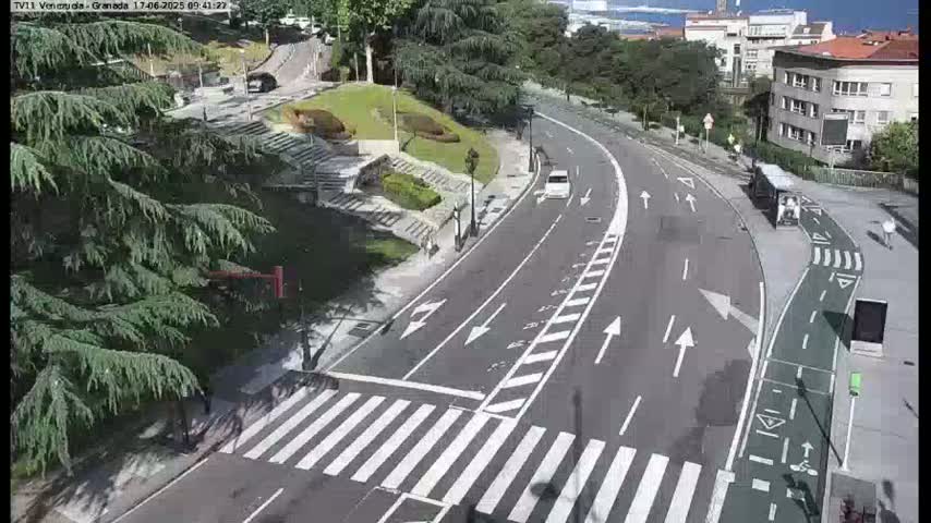 A sunny day shows a winding road with a crosswalk and bike lane, curving through a landscaped area with buildings and greenery visible in the background.