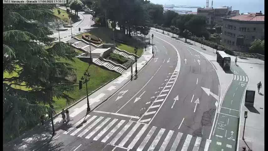 A paved road curves through a park-like setting on a sunny day, with pedestrian crosswalks and a bike lane visible, along with stairs leading up a grassy hillside and some buildings in the distance.