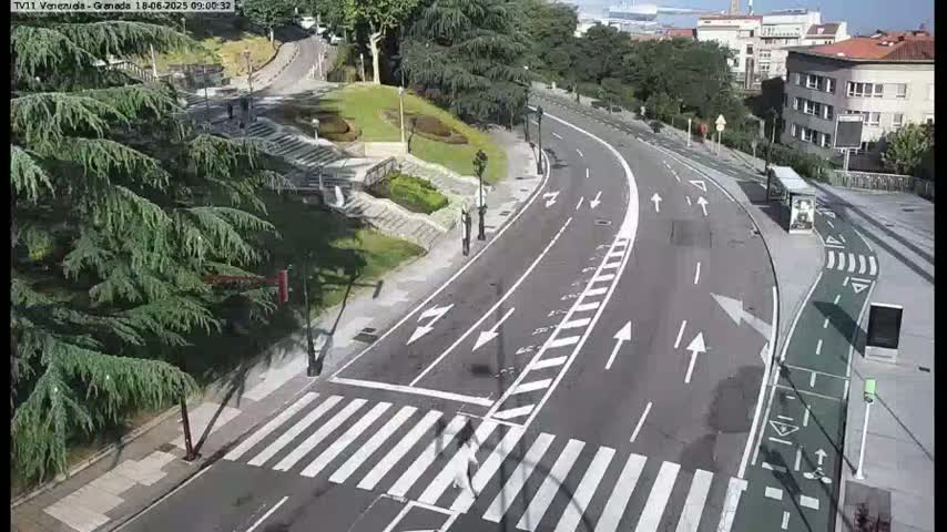 A curving road with crosswalks and a bike lane is seen on a sunny day, next to a grassy area with trees and buildings in the background.