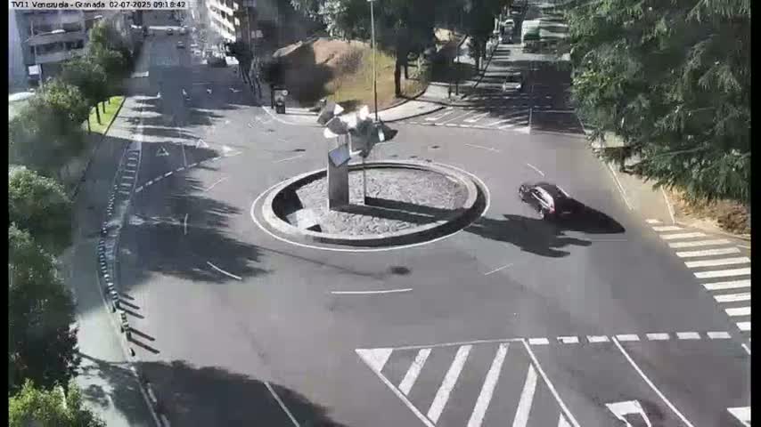A sunny overhead view of a traffic circle with a modern sculpture in the center, surrounded by roads and parked cars.