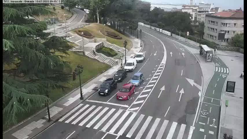 Several cars are driving on a curved road that passes a pedestrian crossing and a staircase leading down to a park-like area on an overcast day.