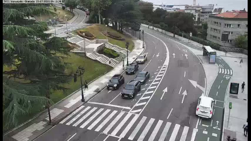 Several dark-colored cars are driving on a curved road that passes by a pedestrian crossing and a park with lush greenery under a partly sunny sky.