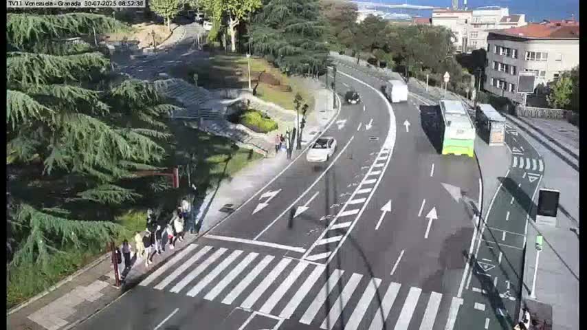 A sunny day shows a roadway curving through a city park with pedestrians crossing a crosswalk, cars driving on the road, and buses stopped at a bus stop.