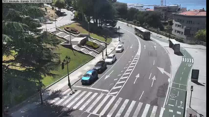 A sunny day shows a roadway curving through a city park with pedestrians crossing a crosswalk, cars driving on the road, and buses stopped at a bus stop.