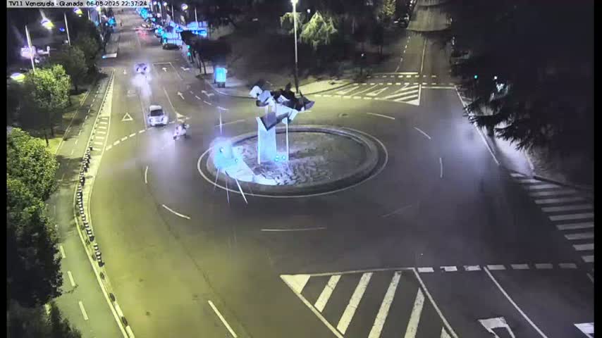 A nighttime, overhead view shows a roundabout with a few vehicles and pedestrians, surrounded by roads and trees under dark conditions.