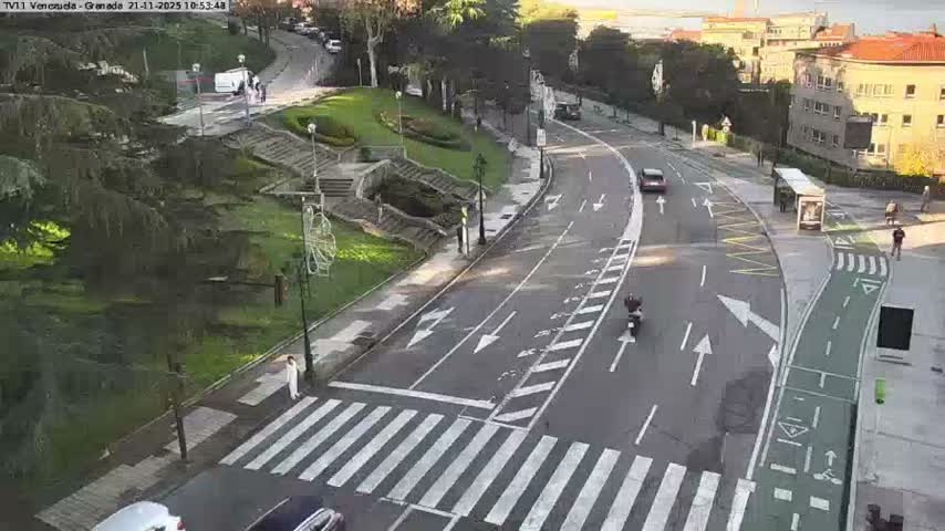 An overhead view reveals a city street with cars, a motorcycle, and pedestrians, featuring a multi-lane road, a dedicated green bike lane, a landscaped park with trees and stairs, and buildings on the right, all under clear, sunny skies.