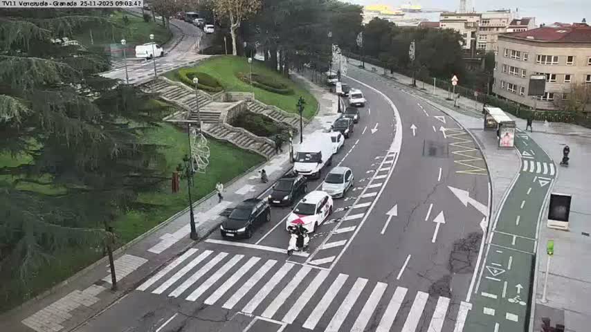 An elevated view captures a curved city street with a line of vehicles, including cars, a van, and a scooter at a crosswalk, a green bike lane, and pedestrians on sidewalks, all set against a backdrop of grassy hills with stone stairs and urban buildings under an overcast sky.