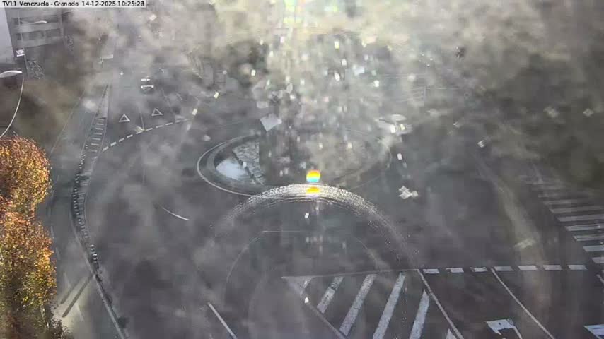 An elevated view captures a curved city street with a line of vehicles, including cars, a van, and a scooter at a crosswalk, a green bike lane, and pedestrians on sidewalks, all set against a backdrop of grassy hills with stone stairs and urban buildings under an overcast sky.