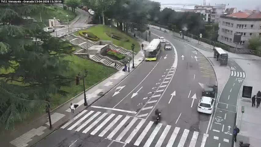 An elevated view captures a curved city street with a line of vehicles, including cars, a van, and a scooter at a crosswalk, a green bike lane, and pedestrians on sidewalks, all set against a backdrop of grassy hills with stone stairs and urban buildings under an overcast sky.