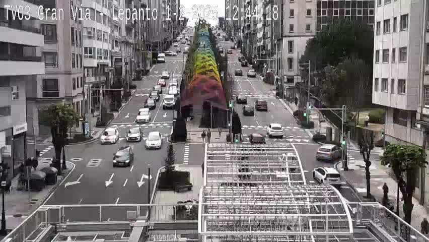 A city street scene on an overcast day shows multiple cars driving on a multi-lane road that passes under a colorful rainbow-like structure.