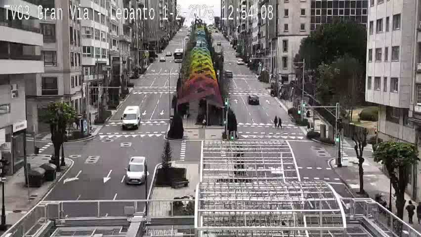 A city street scene on an overcast day shows several cars driving along a wide road with buildings lining both sides and a colorful structure in the center.