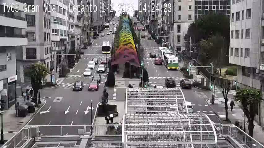 A city street scene on a partly cloudy day shows several cars and buses driving past a large, colorful structure in the middle of the road, with buildings lining either side of the street.