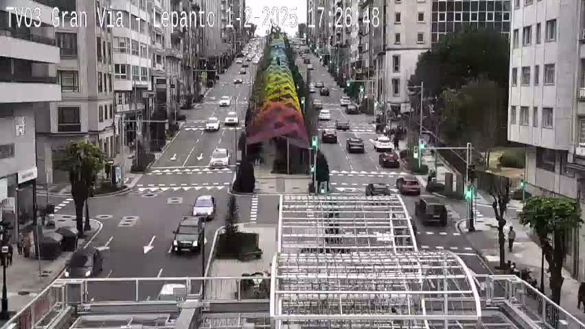 A city street scene with overcast skies shows cars driving on a multi-lane road that intersects with another road, and a rainbow-colored canopy in the center of the road.