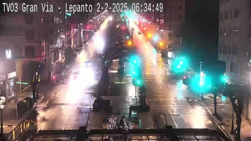 A rain-slicked city street at night shows vehicles driving past traffic signals and buildings.