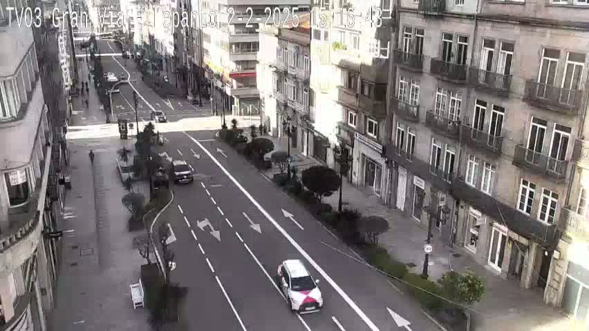 A city street scene on a sunny day shows several cars driving down a wide road lined with multi-story buildings and trees.