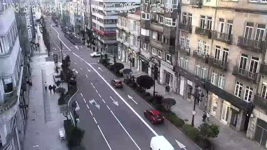 A city street scene on an overcast day shows several cars driving down a multi-lane road that runs between tall buildings.