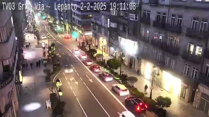 A nighttime, city street scene shows multiple cars driving on a multi-lane road lined with buildings, and pedestrians walking on the sidewalk.