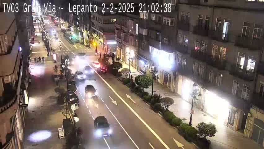 A nighttime, high-angle view of a city street with several cars driving on a two-lane road, lined with buildings that appear to be shops or restaurants.
