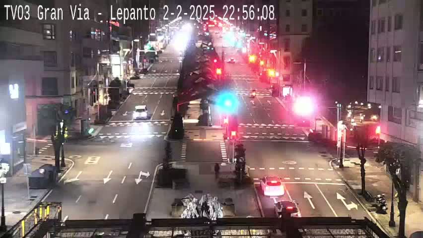 A nighttime, street-level view of a city intersection with several cars and traffic signals, under clear skies.