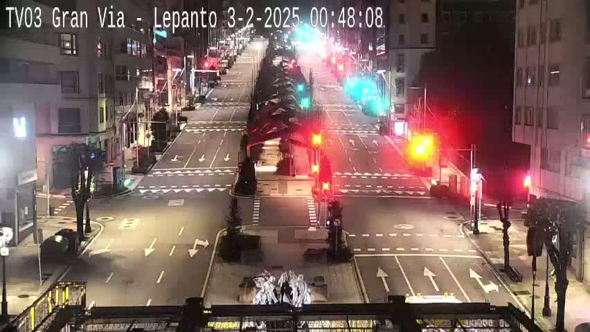 A mostly empty, well-lit city intersection at night with traffic lights and a few cars.