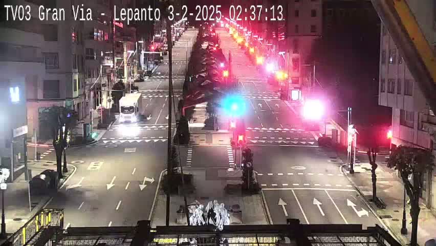 A nighttime, high-angle view of a city intersection with several vehicles and pedestrians, illuminated by streetlights and traffic signals.
