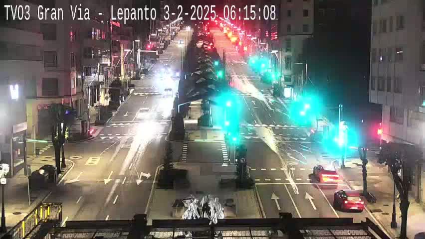 A nighttime, high-angle view of a city intersection with several cars and traffic signals, showing light vehicle traffic on a wet street.