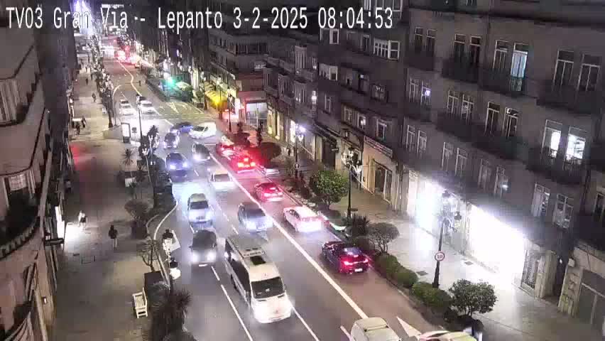 A nighttime, city street scene shows a multi-lane road with moderate traffic flow, lined with buildings and some pedestrians.