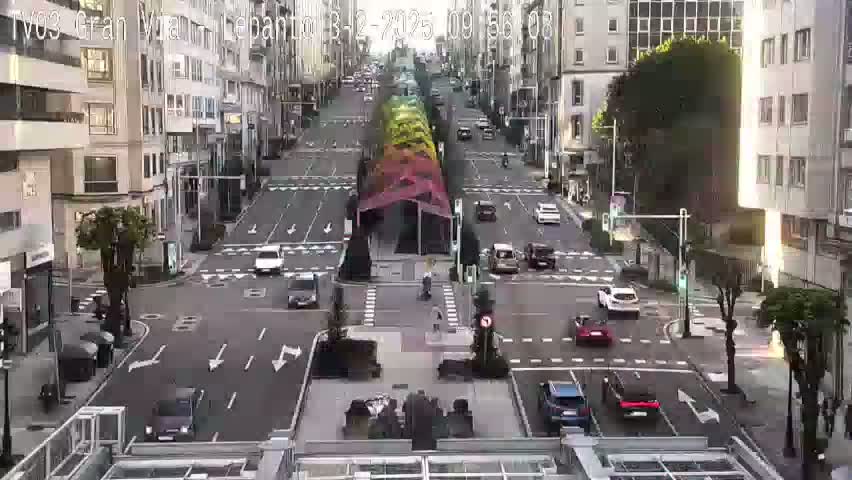 A brightly colored rainbow-like structure is situated in the center of a city intersection where several cars are driving on a sunny day.