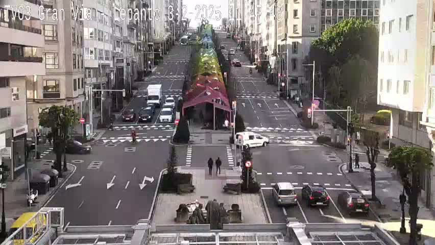 A city street scene with several cars at an intersection, under a mostly clear sky, features a colorful canopy structure in the center of the road.