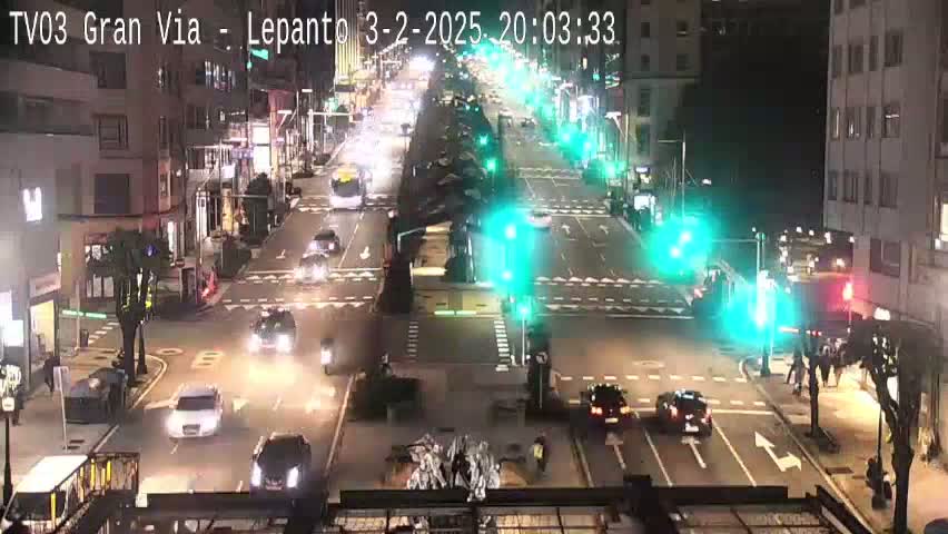 A nighttime, city street scene shows a multi-lane road intersection with numerous cars and pedestrians, under clear skies.