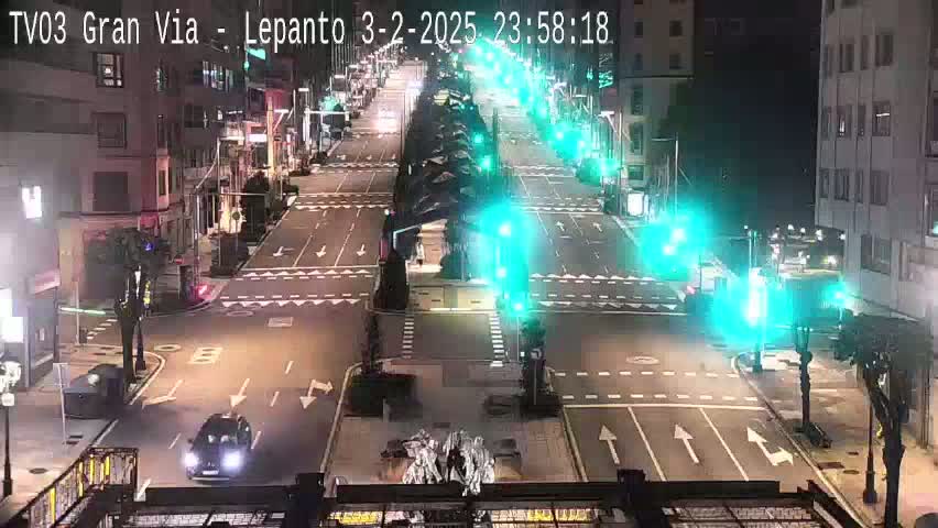 A nighttime, city street scene shows a mostly empty intersection with traffic signals, a few vehicles, and buildings lining the street.