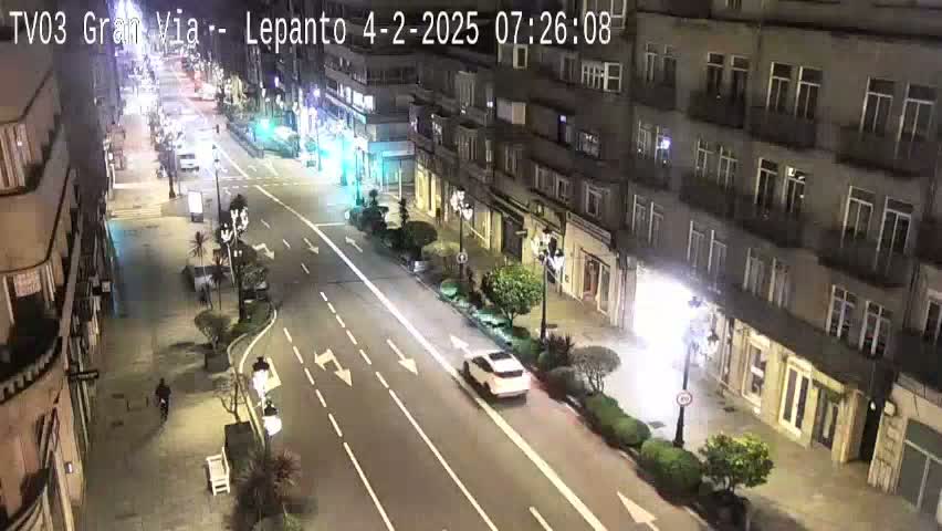 A nighttime, street-level view shows a city street with cars driving on a multi-lane road that's flanked by buildings and small landscaping elements.