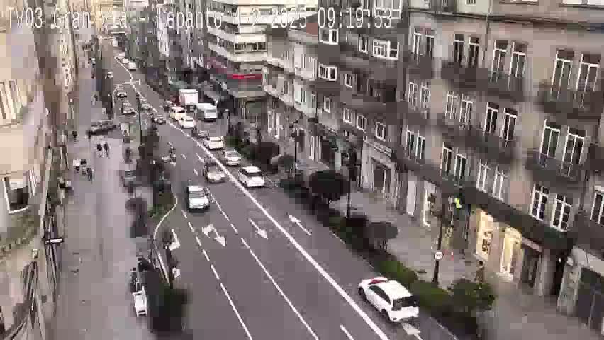 A high-angle view shows a city street with several cars driving in both directions, pedestrians on the sidewalk, and multi-story buildings lining the street on a seemingly overcast day.