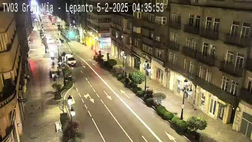 A nighttime street scene shows a multi-lane road with sparse traffic, lined with buildings on either side and sparsely planted trees.