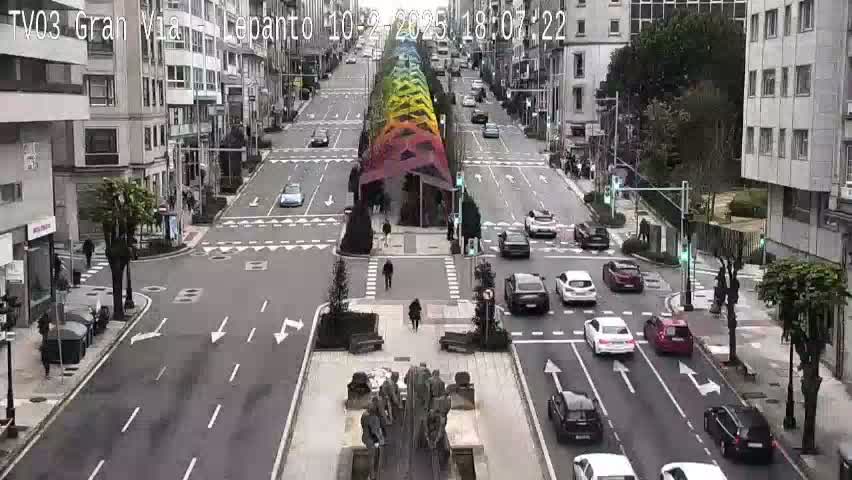 A city street scene on an overcast day shows multiple cars at an intersection near a colorful structure in the middle of the road.