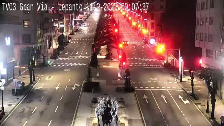 A mostly empty city street at night, with some traffic lights, and a sculpture in the center of the intersection.