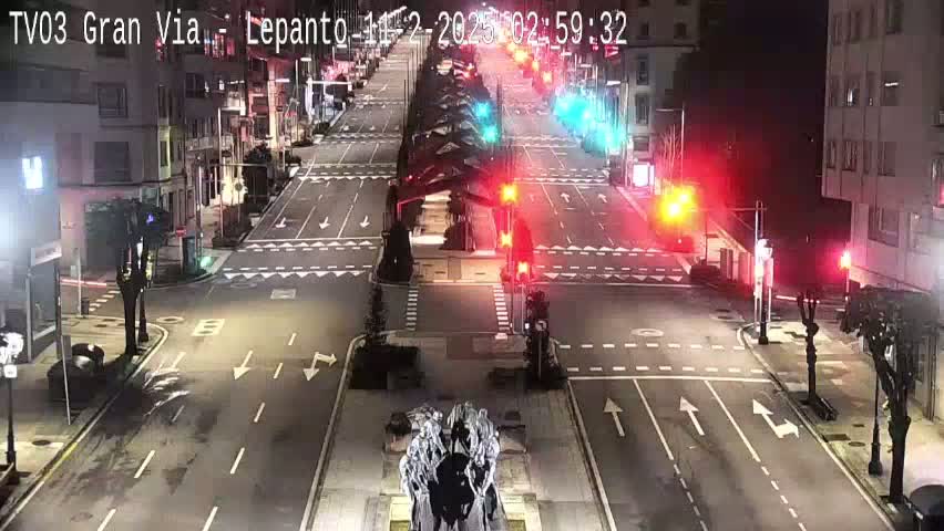 A mostly empty, multi-lane road intersection at night, with traffic lights and buildings lining the streets.