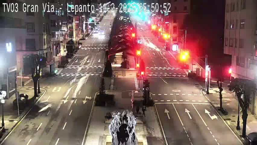 A nighttime, high-angle view of a mostly empty city intersection with traffic lights, buildings lining the streets, and a statue in the center.