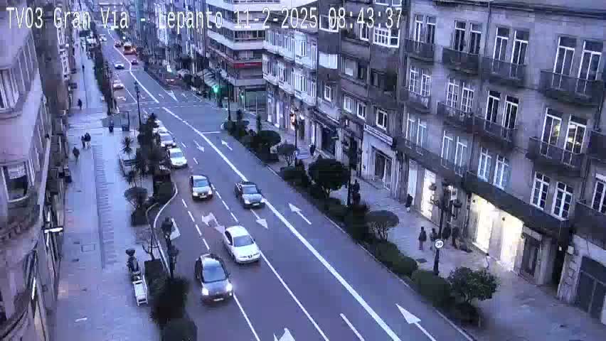 A city street with several cars driving on a roadway lined with buildings under a twilight sky.