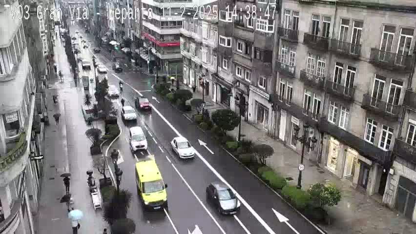 A rainy city street scene shows several cars driving past multi-story buildings with pedestrians walking along the sidewalk.