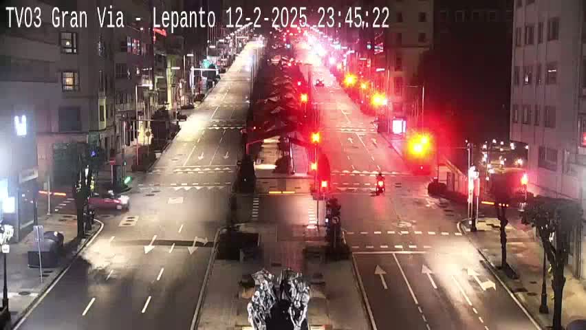 A mostly empty, multi-lane city street at night is illuminated by streetlights and the red lights of traffic signals.