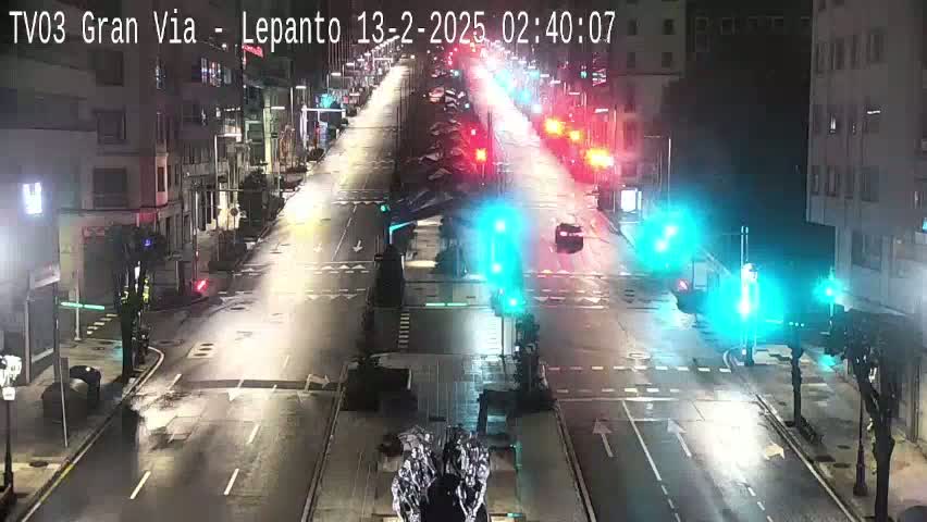 A nighttime, wet city street scene shows several cars driving through a multi-lane intersection with traffic signals.
