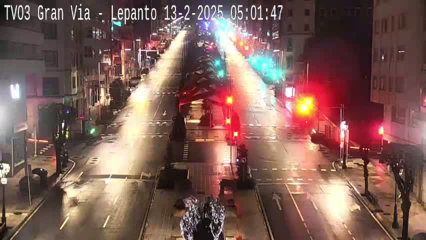 A wet, nighttime city street scene shows several vehicles driving on a multi-lane road with traffic lights and buildings lining both sides.