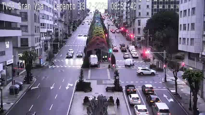 A city street scene shows several cars driving on a multi-lane road that intersects with another road;  a large, colorful, vertical structure is located in the center of the main road; the weather appears to be overcast.