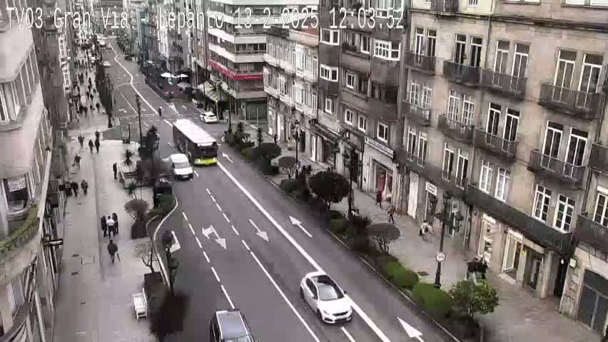 A city street scene shows several vehicles, including buses and cars, traveling on a paved road lined with multi-story buildings; pedestrians are walking on sidewalks.