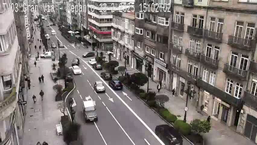 A city street scene on an overcast day shows several vehicles moving along a multi-lane road between tall buildings and pedestrians walking on sidewalks.