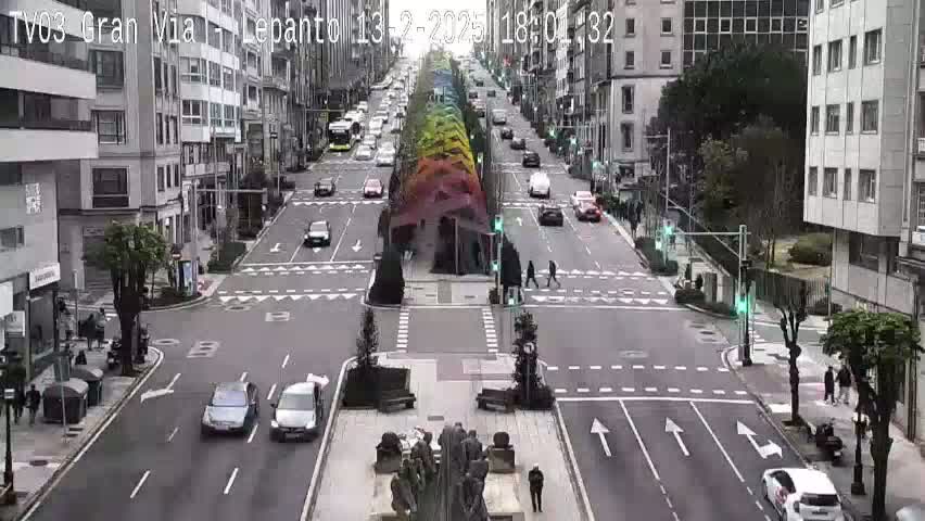 A sunny day shows a city street intersection with several cars and pedestrians, and a rainbow-colored structure in the center of the street.