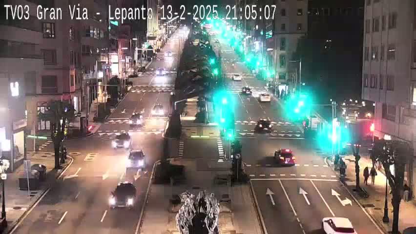 A nighttime street scene shows several cars moving through a city intersection under streetlights, with some pedestrians on the sidewalk.